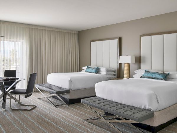 The image shows a hotel room featuring two beds, a small table with chairs, a lamp, and large windows with curtains.