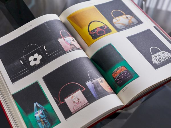 The image shows an open book with pages displaying various handbag designs on a glossy table.