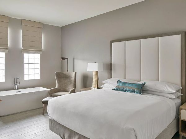 The image shows a modern bedroom with a large bed, a chair, two lamps, a bathtub, and windows, all in a neutral color scheme.