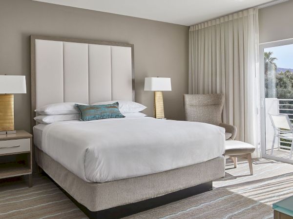 A modern bedroom with a large bed, nightstands with lamps, an armchair, and sheer curtains, overlooking a balcony with a view.