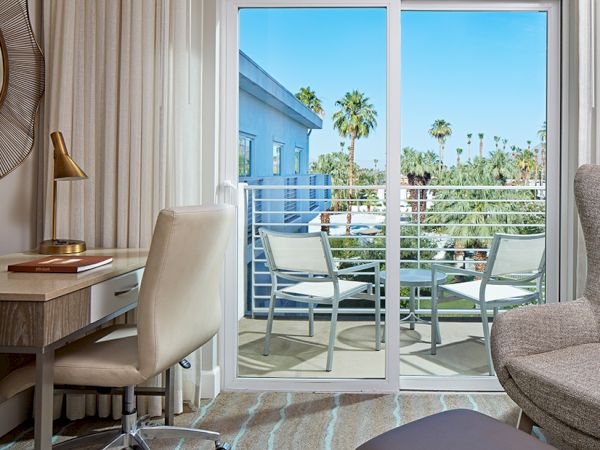 A modern hotel room with a desk, lamp, and chair, leads to a balcony with outdoor seating. Palm trees are visible beyond the sliding glass door.