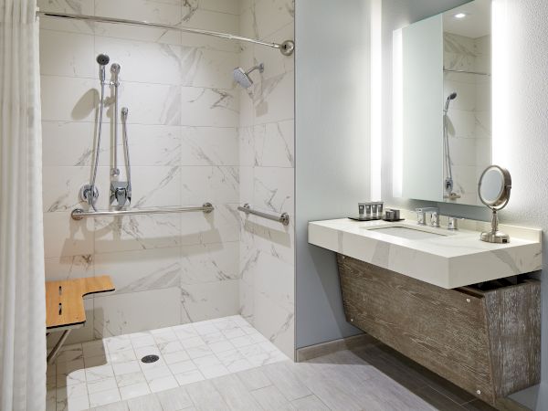 A modern bathroom with a walk-in shower, grab bars, and a wooden bench. Beside it is a vanity with a mirror and a light above.