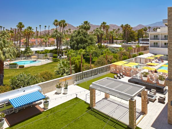 This image shows a sunny outdoor area with palm trees, a pool, lounge areas, and seating, set against a backdrop of mountains and clear skies.