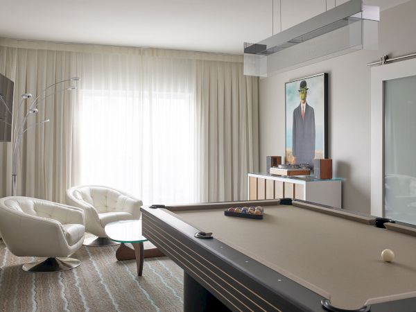 The image shows a modern room with a pool table, white armchairs, a floor lamp, wall art, and a side table against sheer curtains.