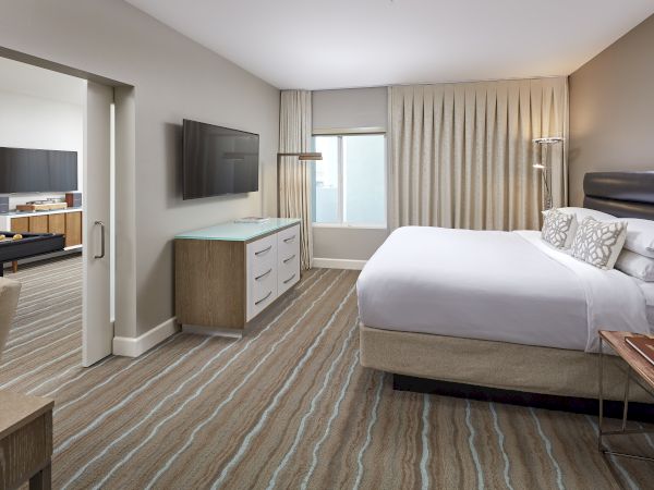A modern hotel room with a bed, dresser, TV, and a connected living area with another TV in the next room, featuring neutral tones.