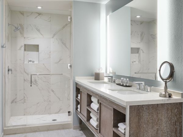 A modern bathroom with a glass shower, marble tiles, a large mirror, a vanity with towels, and a small round mirror on the counter.