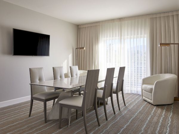 A modern dining room with a glass table, six chairs, a wall-mounted TV, an armchair, and large curtains by a window complete the scene.