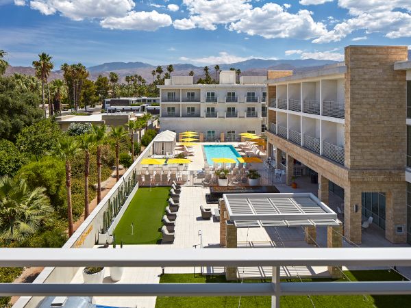 A modern hotel with a central pool, surrounded by palm trees and mountains, under a partly cloudy sky, creating a relaxing atmosphere.