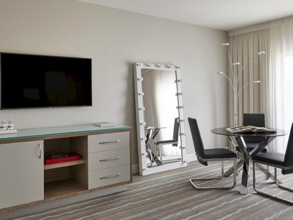 The image shows a modern hotel room with a wall-mounted TV, a large mirror, a dining table with chairs, and sheer curtains.