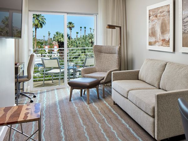 A modern hotel room with a sofa, armchair, desk, and large window showing a balcony with palm trees outside. Art hangs on the walls.