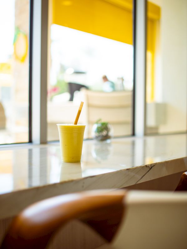 A yellow cup with a straw sits on a bright counter by a sunny window, a cozy cafe vibe in soft focus.