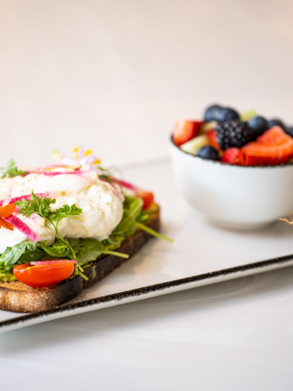 A plated open-face sandwich with greens, tomato, avocado, and a sunny-side egg beside a bowl of fresh berries, on a rectangular tray.