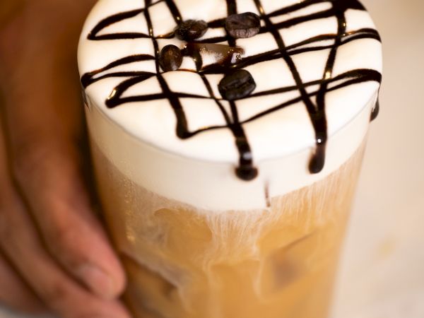 A hand holds an iced coffee topped with foam, drizzled with chocolate syrup in a lattice pattern, and garnished with coffee beans.