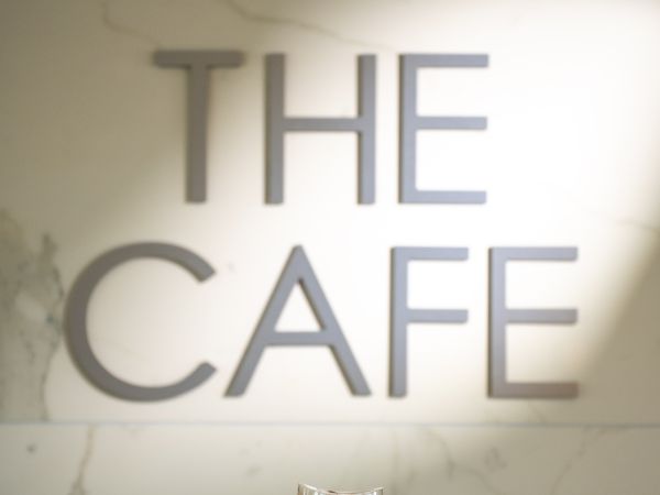 A hand holds an iced coffee with whipped cream in front of a wall with the text "THE CAFE" illuminated by light.
