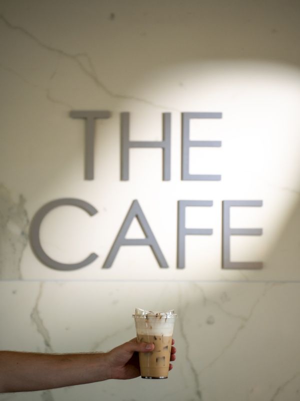 A hand holds a cup of coffee in front of a wall that reads &ldquo;THE CAFE&rdquo; in large letters.