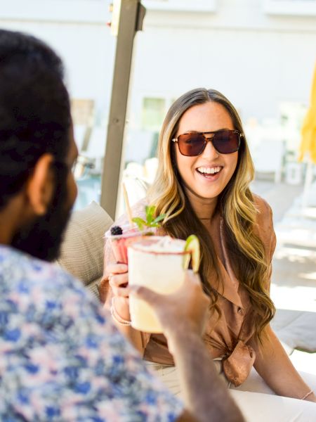 Two people are sitting outdoors by a pool, smiling and toasting with beverages.