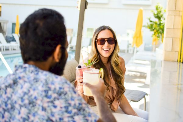 Two people are sitting outdoors by a pool, smiling and toasting with beverages.