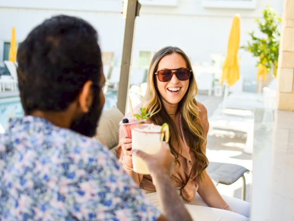 Two people are sitting outdoors by a pool, smiling and toasting with beverages.