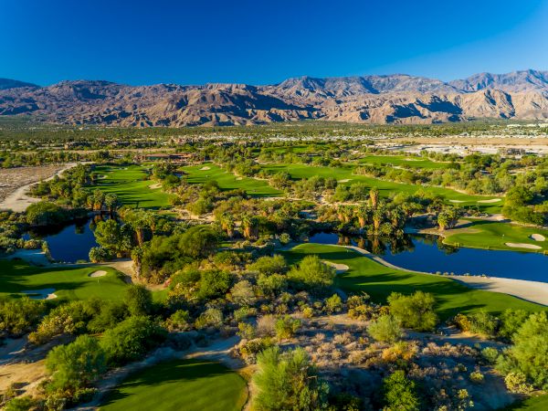 Aerial view of a lush golf course with ponds, surrounded by desert landscape and mountains under a clear blue sky.