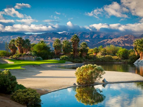 The image shows a scenic golf course with palm trees, a sand trap, a pond, and mountains in the background under a partly cloudy sky.