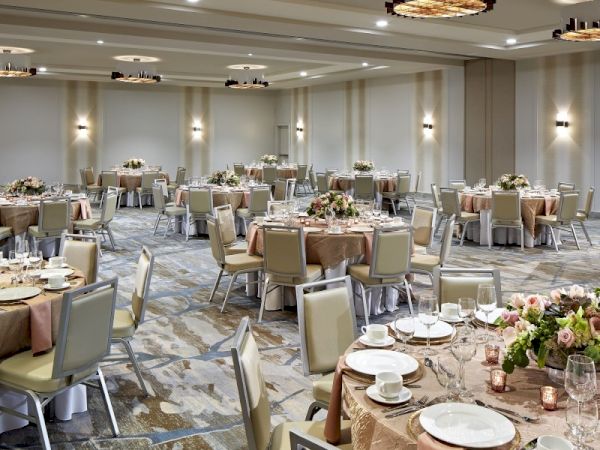 The image shows an elegantly decorated banquet hall set up with round tables, chairs, floral centerpieces, and tableware, ready for an event.