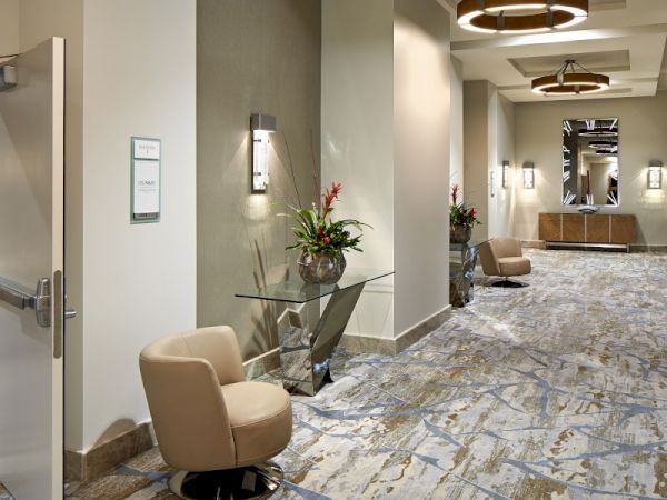 A hotel hallway with stylish decor, featuring a chair, a side table with flowers, wall lights, and patterned carpeting, creating an inviting ambiance.