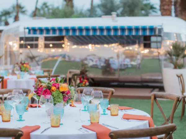 An outdoor setup with tables, vibrant floral centerpieces, colorful glassware, and a retro trailer backdrop amidst palm trees and string lights.