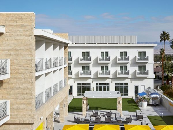 The image shows a modern hotel with multiple stories, featuring a courtyard with green lawns, yellow umbrellas, and outdoor seating areas.