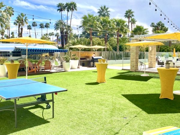 Outdoor area with green turf, tables with yellow covers, umbrellas, a ping pong table, palm trees, and string lights in the background.