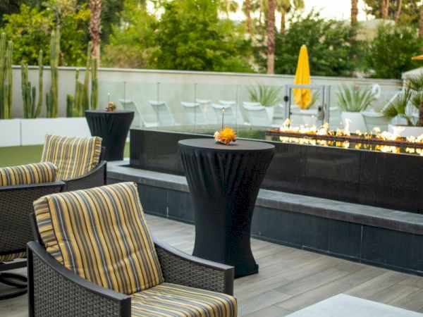 The image shows an outdoor patio with striped cushioned chairs, a black pedestal table, and a fire pit, surrounded by greenery and palm trees.