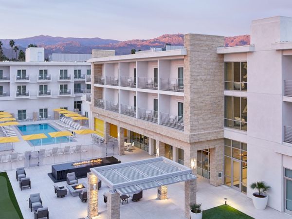 A modern hotel with a pool, outdoor seating area, and a pergola on a green lawn, set against a mountain backdrop under a pastel sky.