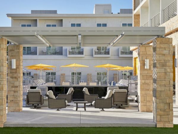 The image shows an outdoor patio with seating, yellow umbrellas, and a modern building in the background, under a clear blue sky.