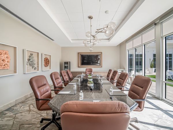 A modern conference room with a long glass table, ten brown leather chairs, large windows, marble floors, and pendant lighting.