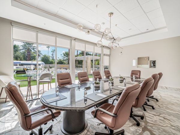 A modern conference room with a long glass table surrounded by brown leather chairs, large windows, marble floor, and a chandelier.