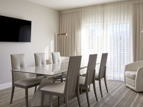 A modern dining room features a glass table, six chairs, a wall-mounted TV, and a large window with sheer curtains.