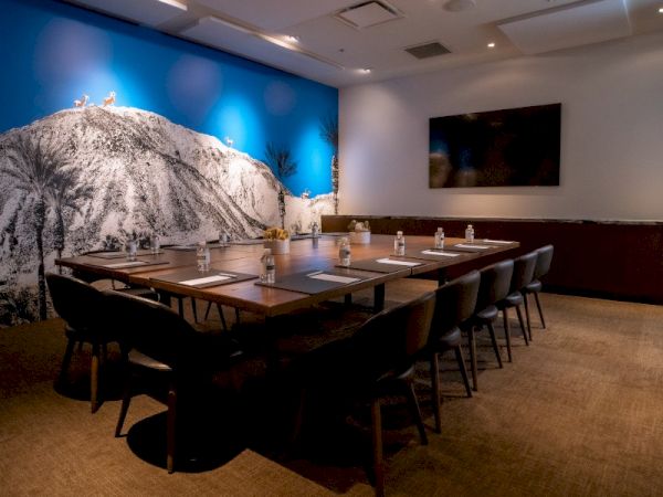 A modern conference room with a long table, chairs, and a large blue mountain mural on the wall. There's also a TV screen.