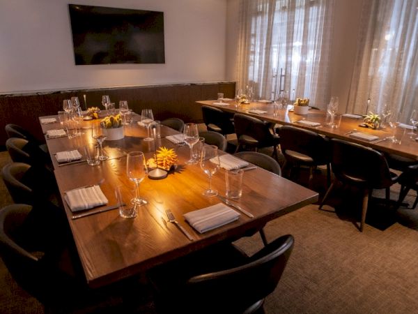 This image shows a dimly lit dining room with a long table set for a meal, featuring wine glasses, napkins, and decorative flowers.