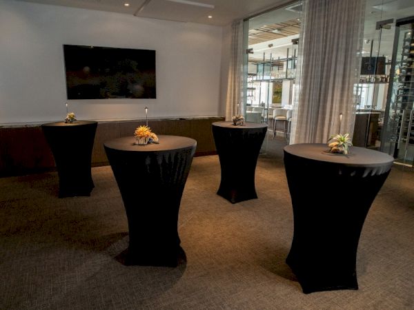 The image shows a dimly lit room with four cocktail tables covered in black cloth, each adorned with a small floral arrangement.