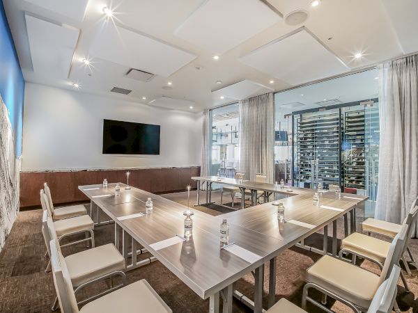 Modern conference room with a long rectangular table, white chairs, a wall-mounted TV, glass windows, and bottled water on the table, ready for a meeting.