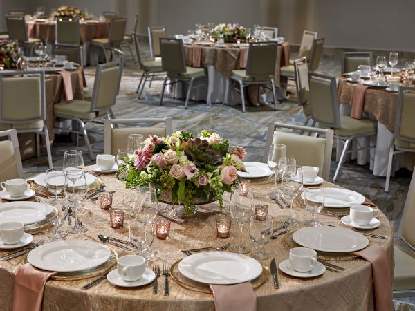 A banquet hall with elegantly set tables, featuring floral centerpieces, plates, glassware, and napkins on beige tablecloths.