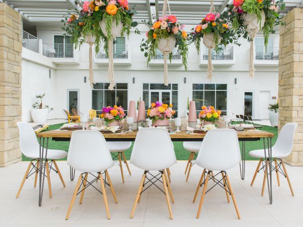 A beautifully set outdoor dining table with white chairs, floral arrangements, and hanging greenery, situated in a patio area.