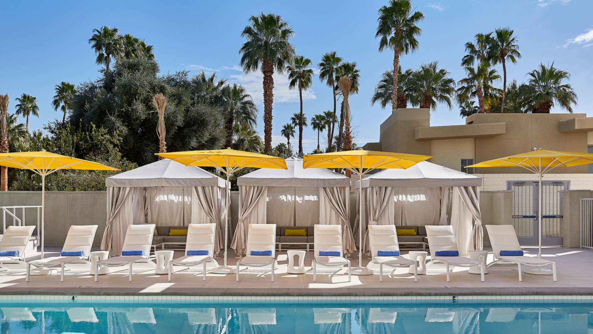 Poolside area with chairs, cabanas, and umbrellas; surrounded by palm trees under a clear blue sky.