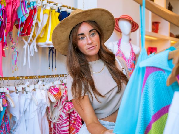 A woman wearing a hat is shopping for clothes in a store with colorful swimsuits displayed on racks.