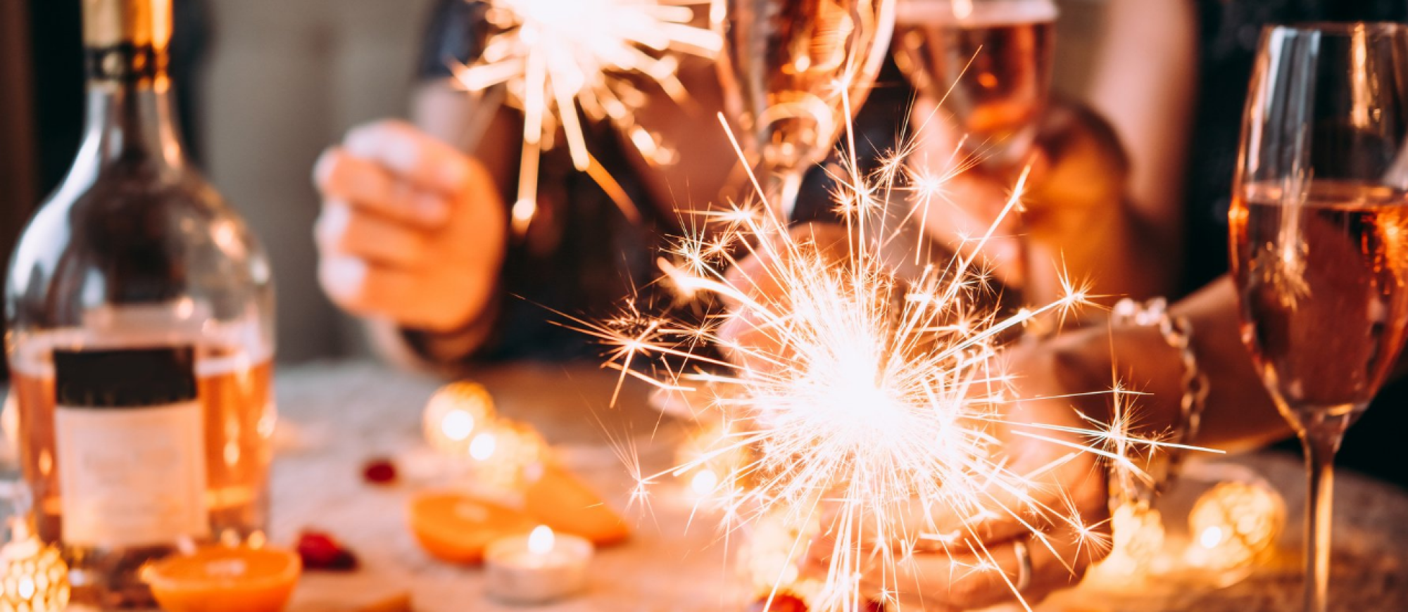 The image shows people holding sparklers and glasses of wine at a festive gathering, with soft lighting and a celebratory atmosphere.