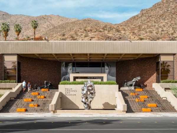 The image shows the exterior of the Palm Springs Art Museum, with steps, sculptures, and a desert mountain backdrop under a blue sky.