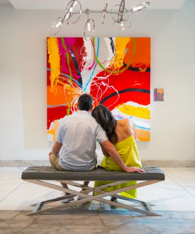 A couple sits on a bench, facing a vibrant abstract painting with bold reds and oranges in a gallery setting.