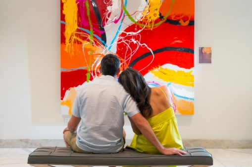 A couple sits on a bench, facing a vibrant abstract painting with bold reds and oranges in a gallery setting.