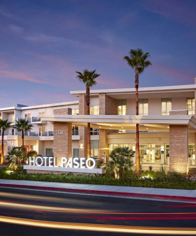 A modern hotel exterior with a lit "HOTEL PASEO" sign, palm trees, and light trails from passing vehicles under a dusk sky.