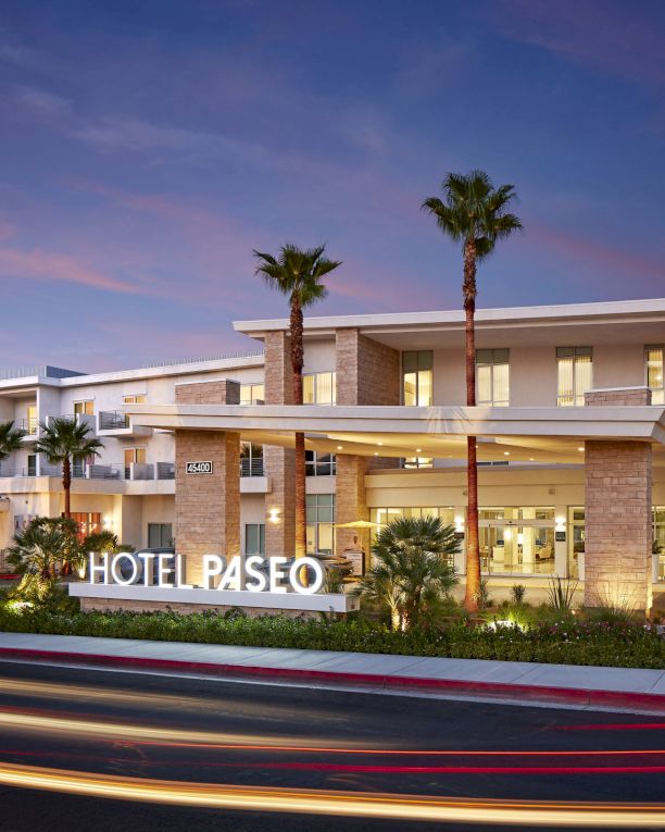 A modern hotel exterior with a lit "HOTEL PASEO" sign, palm trees, and light trails from passing vehicles under a dusk sky.