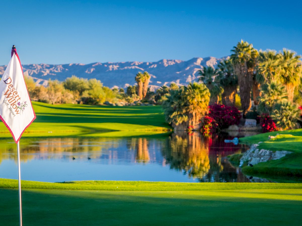 A golf course with a flag labeled "Desert Willow Golf Resort," surrounded by palm trees, a pond, and mountains in the background.